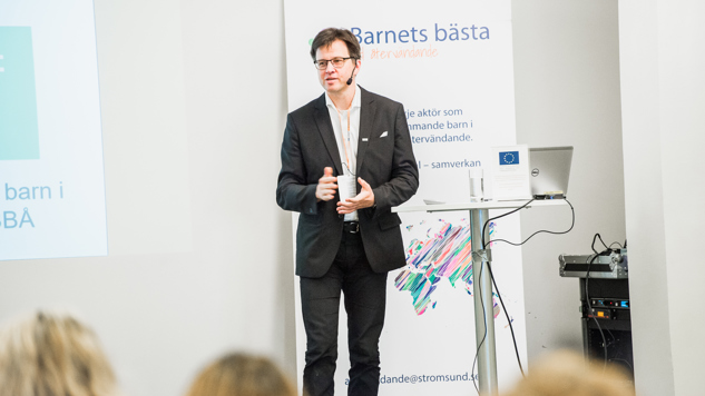 Henrik Malm Lindberg står på en scen och håller i en presentation med en roll-up i bakgrunden där det står "Barnets bästa vid återvändande". 