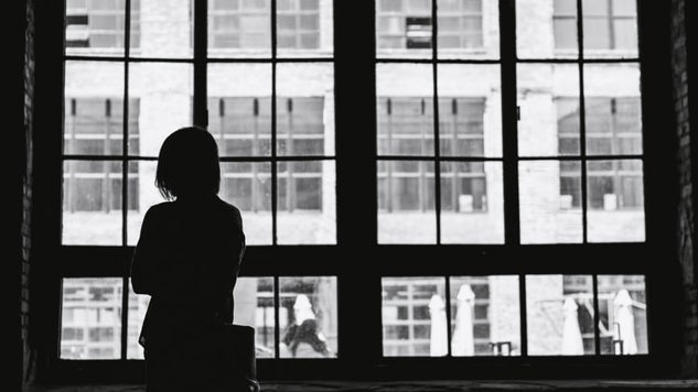 En svart/vit bild av en kvinna som står vid ett fönster. Bild: Unsplash.