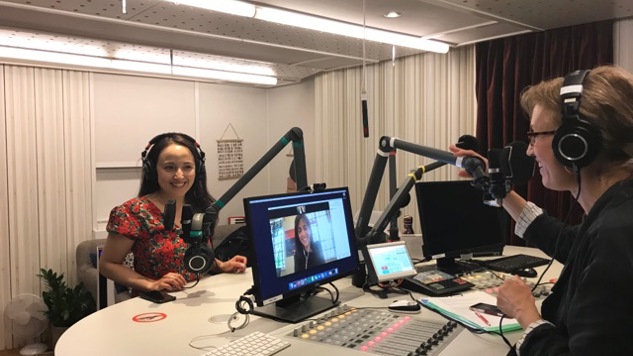 The host talks to a guest sitting in front of her and a guest participating digitally and who is visible on the computer screen.