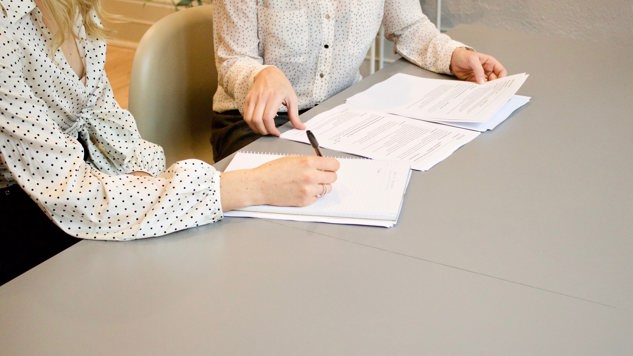 En kvinna som signerar ett papper bredvid en annan kvinna som håller i en bunt papper. Bild: Unsplash.